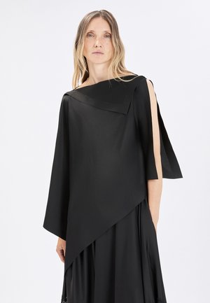 Donna con capelli biondi lunghi che indossa una maglia nera asimmetrica con manica ampia e spalla scoperta su sfondo bianco uniforme.