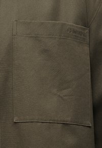 Τσεπάκι από ελαιί πράσινο ύφασμα με ορατές ραφές στο ρούχο, με μικρό ανάγλυφο λογότυπο Calvin Klein κοντά στην πάνω δεξιά γωνία.