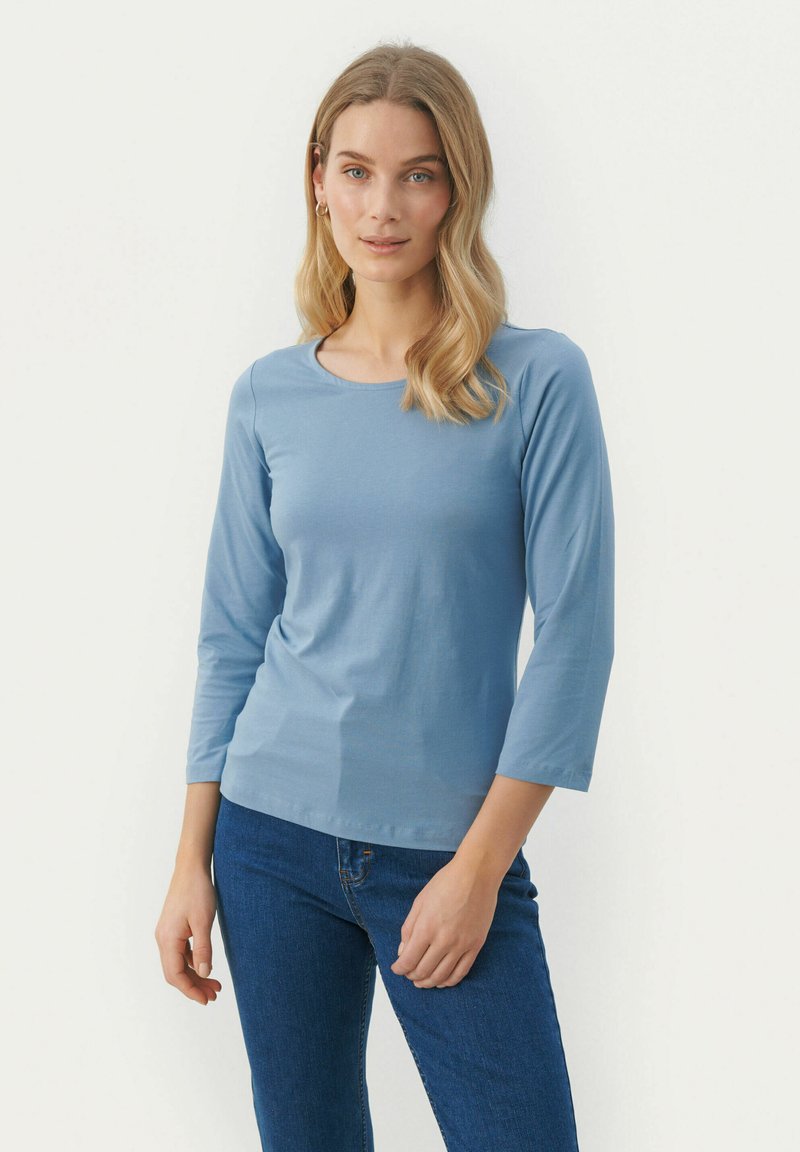Camicia a maniche lunghe azzurro chiaro con scollatura rotonda, texture morbida e forma aderente, abbinata a jeans blu scuro.