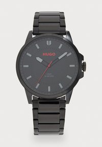 Zwart roestvrijstalen horloge met een ronde wijzerplaat, matte zwarte wijzerplaat, rode accenten, zilveren uurmarkeringen en een drievoudige schakelband. Waterbestendig tot 30 meter.