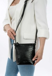 Kvinna som bär en vit blazer och blå jeans, håller en liten svart lädercrossbodyväska med krokodilmönster.