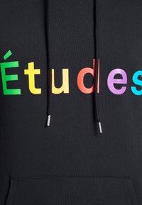 Czarna bluza z kapturem z napisem "Études" w kolorowych literach: zielonym, żółtym, czerwonym, purpurowym i niebieskim. Zawiera sznurki ze srebrnymi metalowymi końcówkami oraz kieszeń na froncie.