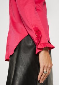 Mano de mujer con anillos de plata cerca de una falda de cuero negra y una blusa roja con mangas con volantes y abertura lateral.