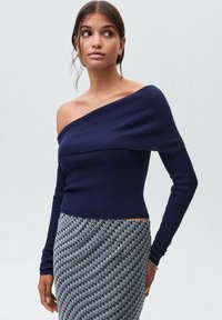 Marine off-the-shoulder ribbet top med lange ærmer, parret med en mønstret nederdel med små geometriske designs i grå og sort.