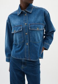 Giubbotto di jeans blu scuro con colletto classico, tasche sul petto, chiusura a bottoni e vestibilità ampia. Tessuto texturizzato con cuciture a contrasto.