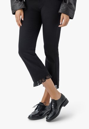 Donna con pantaloni neri a tre quarti con bordo arricciato a pois, giacca di pelle nera e lucide scarpe stringate brogue nere.