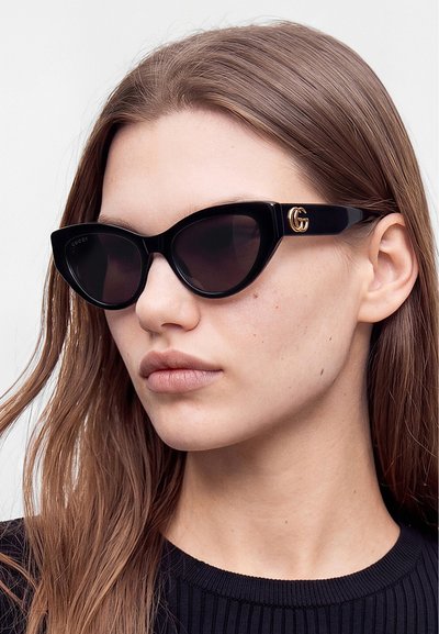 Junge Frau mit langen braunen Haaren, die schwarze Cat-Eye-Sonnenbrillen mit goldenem Gucci-Logo an den Bügeln und ein schwarzes, geripptes Top trägt.