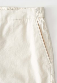 Beige cotton shorts with an elastic waistband, featuring a side pocket and smooth texture. Stitching is visible along the waistband and pocket.