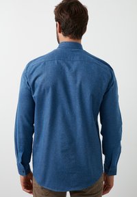 Camisa de hombre de manga larga en una tela azul texturizada. Presenta un cuello estándar, puños con botones y un dobladillo ligeramente curvado.