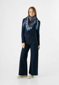 Écharpe à motif bleu nouée au cou, chemise à manches longues bleu foncé, jeans en denim taille haute à jambes larges, et baskets beiges. Une tenue simple et décontractée.