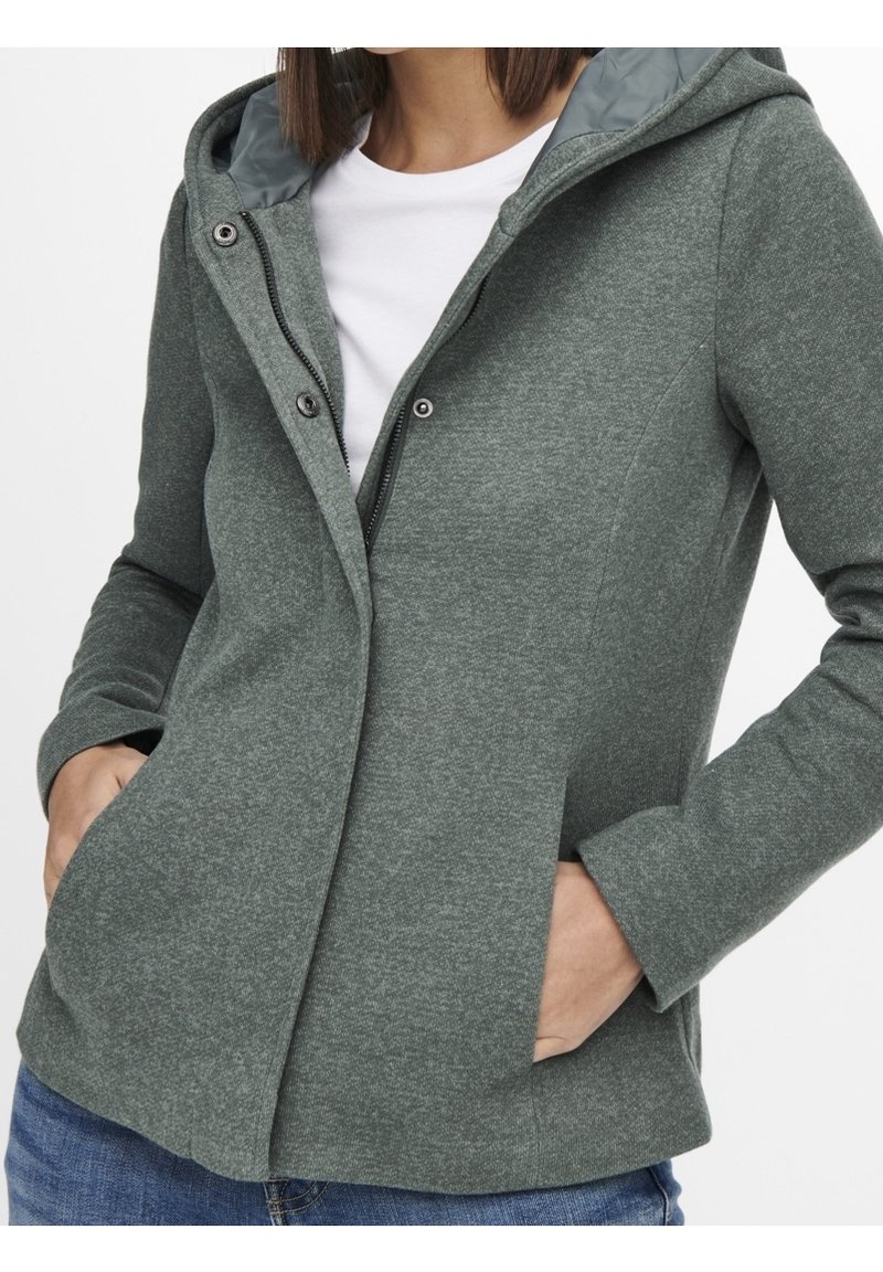 Veste à capuche verte avec fermeture éclair et boutons-pression portée sur une chemise blanche, mains dans les poches et jean bleu visible.