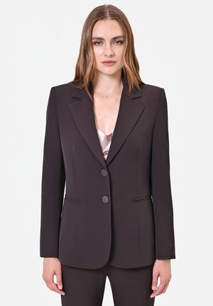 Femme aux cheveux châtain clair portant un blazer marron foncé cintré avec deux boutons et un pantalon assorti sur un fond clair uni.