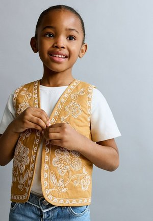 Jong meisje met gevlochten haar lacht terwijl ze een mosterdkleurig vest met witte bloemenpatronen over een wit shirt dichtknopen.