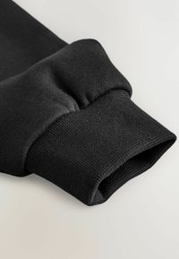 Czarny rękaw od bluzy z ribbowanym mankietem, z gładką teksturą i czystymi szwami, odzwierciedlający dopasowany krój i prosty design.