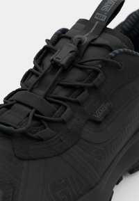 Black hiking shoe featuring textured synthetic material, elastic laces, locking toggle, and a patterned inner lining. Vans branding visible.