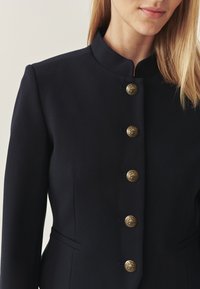 Woman wearing a tailored black blazer with a stand collar and five ornate gold buttons down the front, with part of her lips and hair visible.