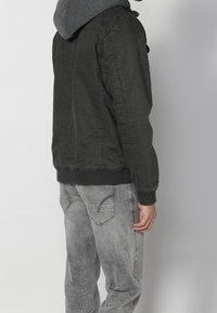 Homme portant une veste vert foncé avec une capuche grise et un jean gris délavé, vu de dos sur un fond clair uni.