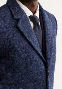 Uomo che indossa un blazer testurizzato blu scuro, una camicia bianca e una cravatta a maglia blu navy su uno sfondo chiaro.