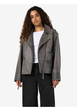 Femme aux longs cheveux bouclés portant une veste en daim grise de style biker sur une chemise blanche et un pantalon large noir, sur un fond blanc.