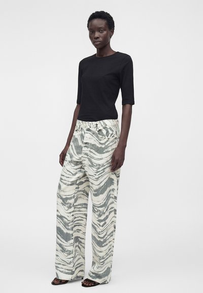 Γυναίκα στέκεται με το πρόσωπο μπροστά φορώντας μαύρο κοντομάνικο τοπ και άνετο λευκό παντελόνι με γκρι αφηρημένα σπειροειδή σχέδια και μαύρα σανδάλια.