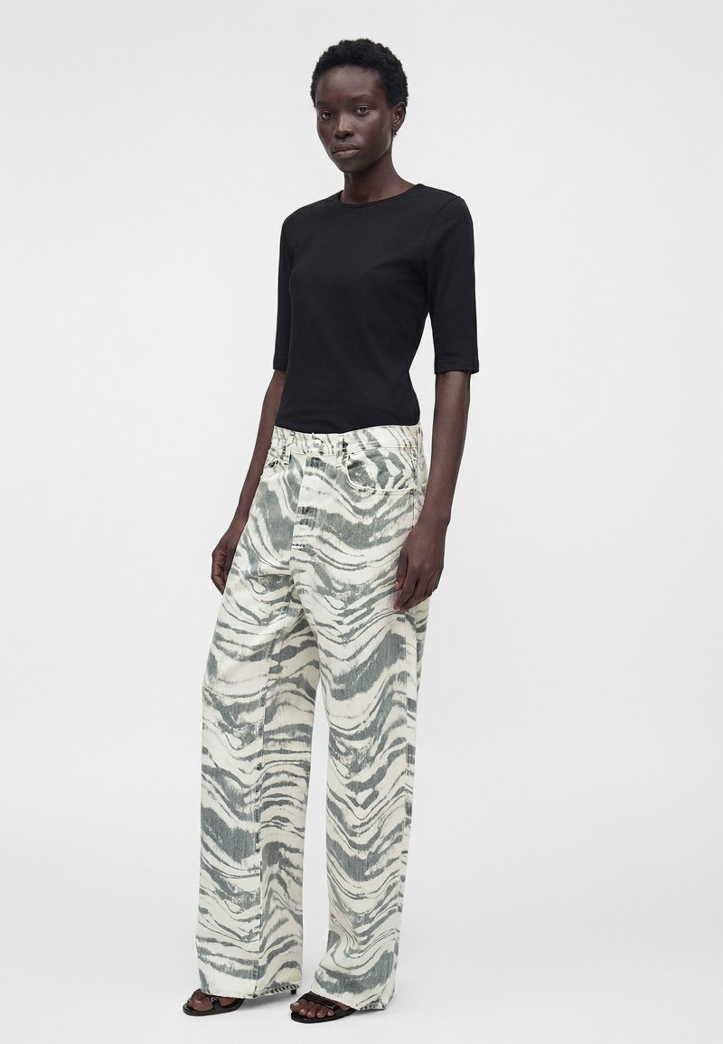 Frau steht nach vorne gewandt und trägt ein schwarzes kurzärmeliges Oberteil sowie lockere weiße Hosen mit grauen abstrakten Wirbelmustern und schwarze Sandalen.