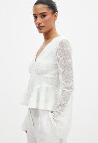 Haut peplum blanc avec broderie florale, décolleté en V et manches longues transparentes. Tissu léger avec une finition douce et texturée.