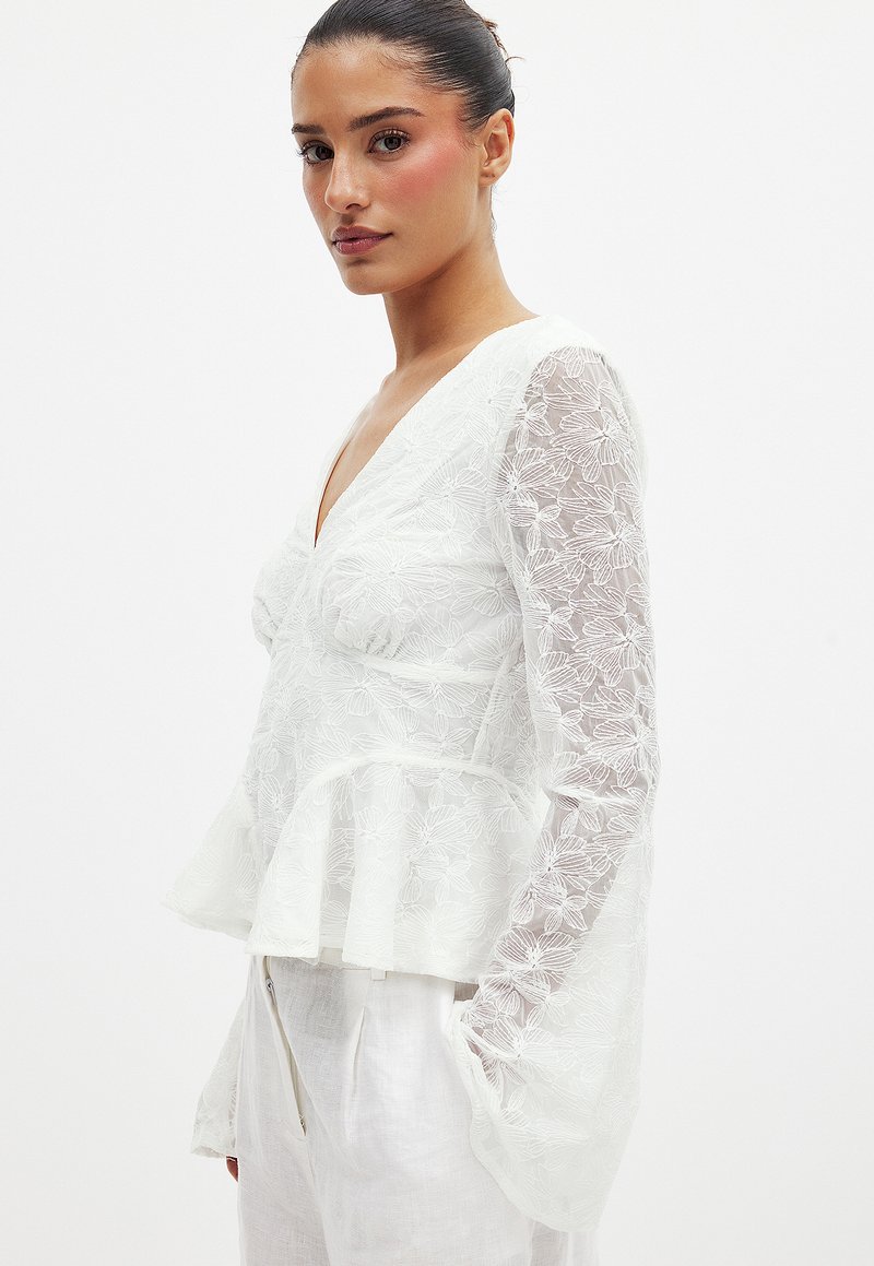 Haut peplum blanc avec broderie florale, décolleté en V et manches longues transparentes. Tissu léger avec une finition douce et texturée.