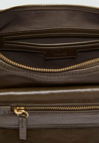 Notranjost rjave usnjene torbice z kontrastnim svetlim oblogo, vsebuje zadržno žep in oznako blagovne znamke. Vidna zlata zadrga.