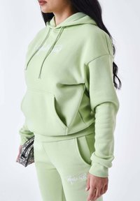 Sudadera verde claro con bolsillo delantero y cordones, con bordado de logo blanco. Combinada con joggers a juego y un bolso texturizado.