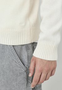 Gros plan d'une main reposant sur le côté, portant un pull à côtes blanc et un pantalon en denim gris avec une poche avant visible.