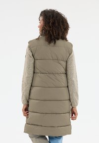 Langer olivgrüner Puffer-Mantel mit einem strukturierten beigefarbenen Pullover darunter. Verfügt über gesteppten Teile und einen hohen Kragen und reicht bis zur Mitte des Oberschenkels.