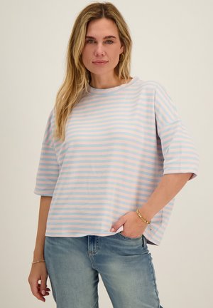 Vrouw met blond haar die een loszittend overhemd met pastelblauwe en roze strepen draagt en lichtblauwe spijkerbroek, poserend met één hand in de zak.