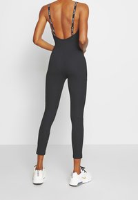 Černý žebrovaný overal s širokým, nízkým výstřihem na zádech a tenkými ramínky s logem. Přiléhavé nohavice končící u kotníků, v kombinaci s bílými teniskami.