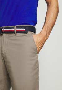Hombre con pantalones beiges y una camisa azul, cinturón azul marino con rayas rojas, blancas y negras, con la mano en el bolsillo de los pantalones.