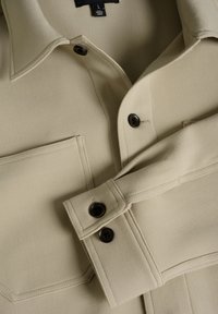 Camicia beige a maniche lunghe con colletto, due tasche sul petto e bottoni scuri. Texture liscia e design strutturato con cuciture visibili.