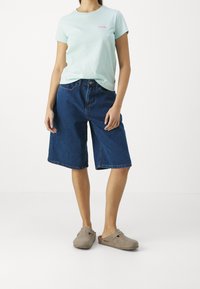 Vaaleansininen puuvillapaita, jossa on vaaleanpunainen logo, yhdistettynä väljiin tummansinsisiin farkkushortseihin. Harmaat liukumattomat kengät, joissa on koristeellinen hihna.