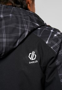 Schwarze Jacke mit grauem Karomuster auf der Kapuze, ausgestattet mit dem weißen "DARE2B"-Logo auf der linken Schulter. Strukturiertes Material, funktionales Design.