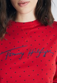 Röd sweatshirt med ett marinblått hjärt-mönster och "Tommy Hilfiger"-logotyp i kursiv stil. Tillverkad av mjukt material med rundad hals.