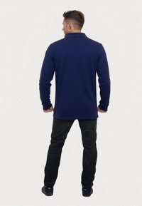 Polo a maniche lunghe di colore blu navy con colletto, abbinato a pantaloni neri e scarpe scure. Il tessuto appare liscio senza motivi visibili.