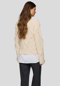 Strickpullover in Creme mit einem strukturierten, fransenbesetzten Design und Diamantmustern. Übereinander getragen mit einem weißen Hemd, kombiniert mit dunkler Hose.