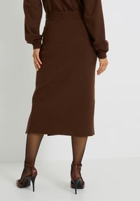 Falda de punto marrón con corte recto, aberturas laterales y una cinturilla ajustada, combinada con zapatos de tacón alto marrones y pant stockings negros transparentes.