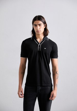 Jeune homme aux cheveux mi-longs portant un polo noir avec des bordures blanches et un jean foncé, se tenant devant un fond gris clair uni.