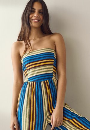 Tânără cu păr lung și închis la culoare, purtând un combinezon fără bretele cu dungi în nuanțe de albastru, galben, maro și crem, stând în fața unui perete simplu.