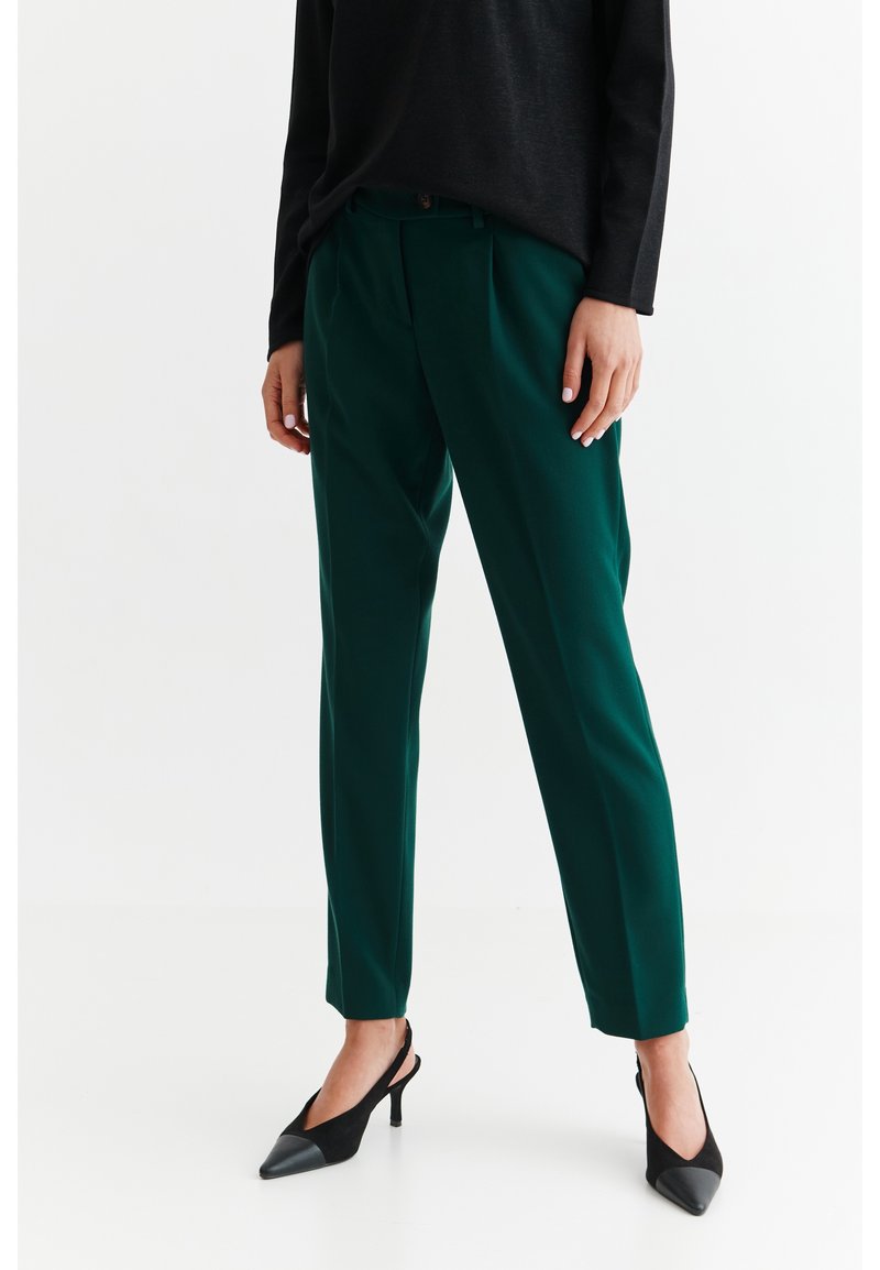 Femme portant un pantalon vert foncé sur mesure, un top noir à manches longues et des talons slingback noirs à bout pointu, debout sur un sol blanc.