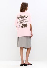 Lyserød oversized t-shirt med fed sort grafisk tekst, parret med en grå nederdel og sorte sandaler med en maroon rem.