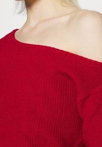 Czerwony sweter z dzianiny z odkrytymi ramionami, o prążkowanej fakturze i szerokim dekolcie. Miękki materiał z widocznymi szwami.