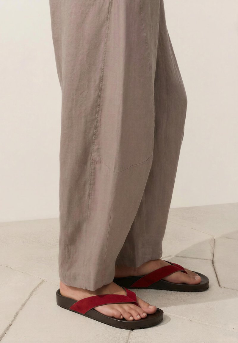 Bas des jambes portant un pantalon beige ample et des sandales en daim rouge avec des semelles noires, debout sur un sol en pierre clair.