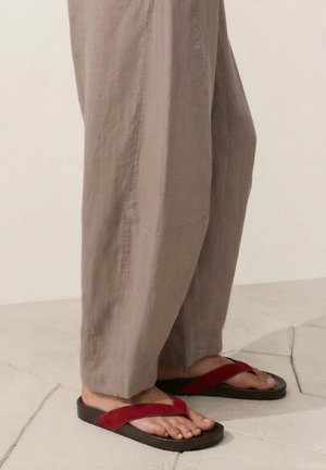 Parte inferiore delle gambe che indossano pantaloni beige larghi e sandali in pelle scamosciata rossi con suole nere in piedi su un pavimento chiaro in pietra.