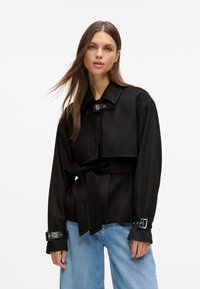 Czarna kurtka z mieszanki wełny z paskiem w talii, dużym kołnierzem i klamrami na mankietach. Gładka tekstura, oversize'owy krój i minimalny design.