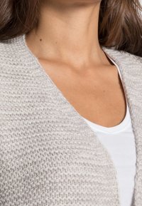 Jasnoszara, dziergana kardigan z teksturowanym wzorem i otwartym przodem, noszony na białą koszulę z dekoltem w serek. Zbliżenie na dekolt i materiał.